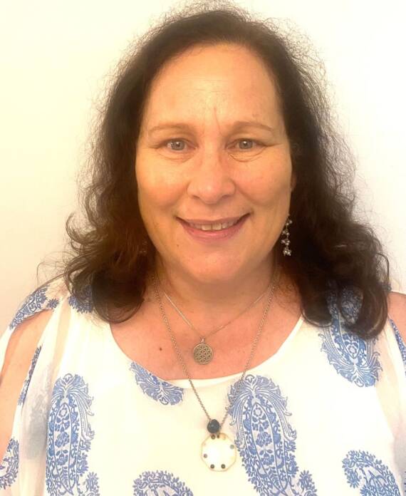 Woman with long brown hair wearing a white and blue patterned top, earrings, and layered necklaces, smiling in front of a plain light background.