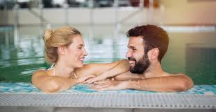 A woman and a man are standing in a swimming pool, facing each other and smiling, with their arms resting on the edge of the pool.