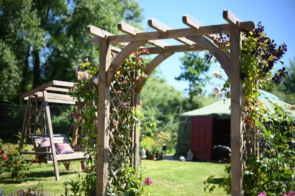 A wooden garden arbour with climbing plants stands in a grassy garden—perfect for relaxing after a massage in Guildford. A wooden swing and a round tent are visible in the background on a sunny day.