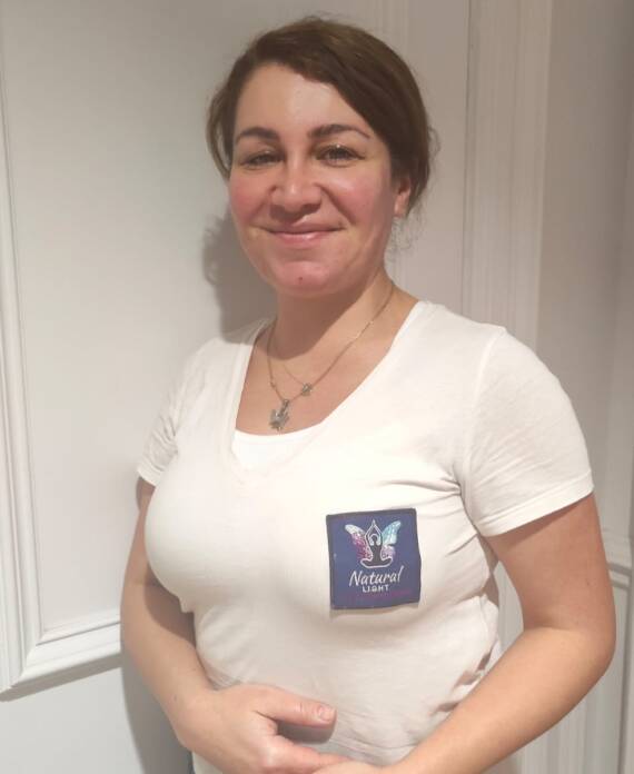 A woman stands indoors wearing a white T-shirt with a butterfly logo and "Natural Lash" text on a badge. She is smiling and looking at the camera.