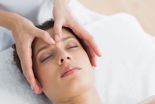 Person lying with eyes closed receiving a facial massage, with hands pressing gently on their forehead, on a white towel.