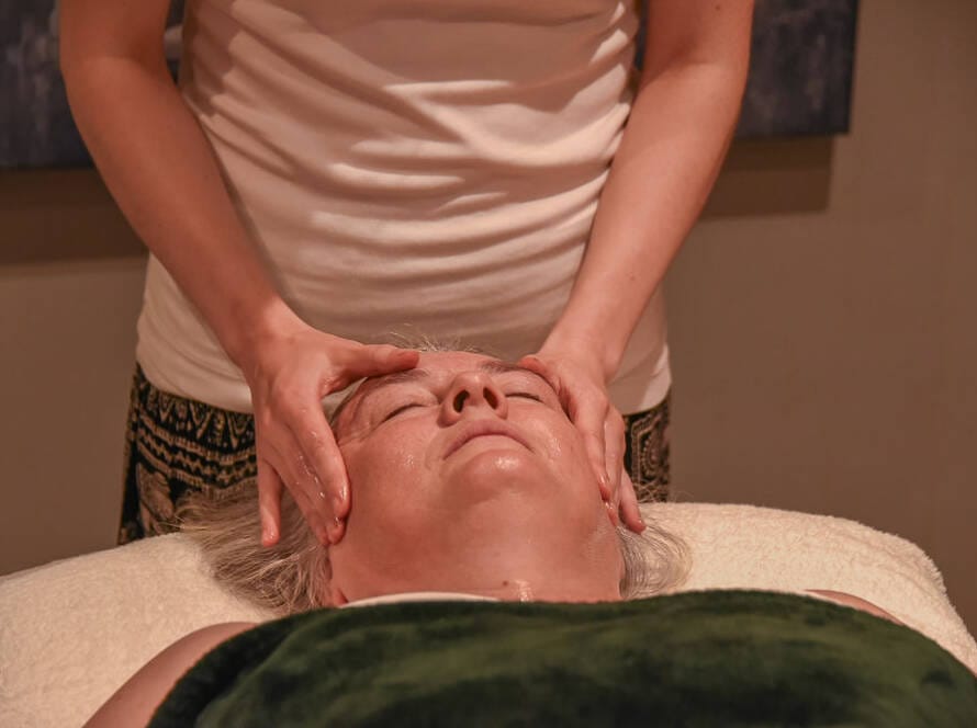 A person lying on a massage table receives a facial massage from another individual using both hands.