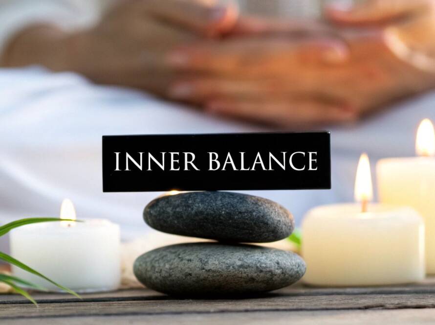 Three smooth stones are stacked below a sign that reads "INNER BALANCE," surrounded by lit candles, green leaves, and a person sitting in the background.