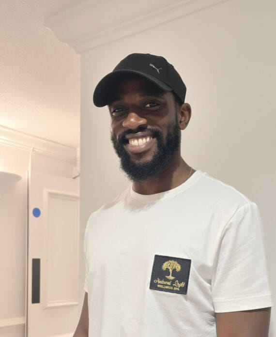 Man wearing a black cap and a white T-shirt with a "Natural Light Wellness" logo stands indoors, smiling at the camera.