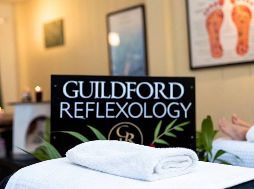 A neatly folded white towel sits on a massage table with a Guildford Reflexology sign in the background and a pair of feet visible to the right.