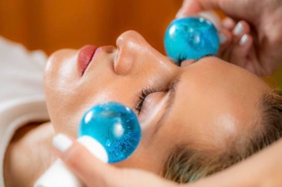 A person receives a facial treatment with two blue glass massage globes applied under their eyes.