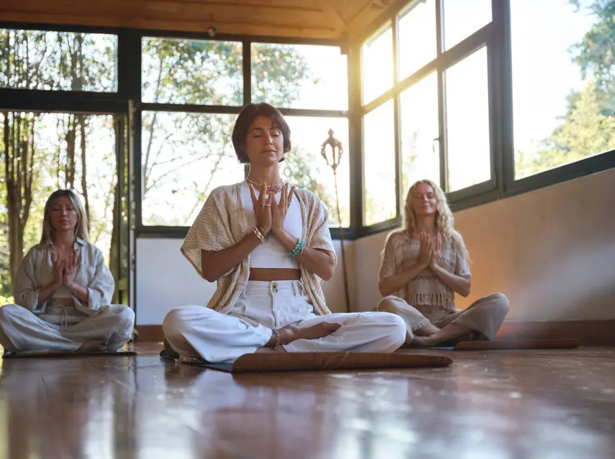 Three women sit cross-legged on yoga mats in a sunlit room, eyes closed and hands in prayer position, meditating.