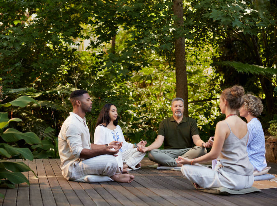 Five people sit cross-legged on a wooden decking outdoors, meditating in a circle surrounded by green trees and plants.