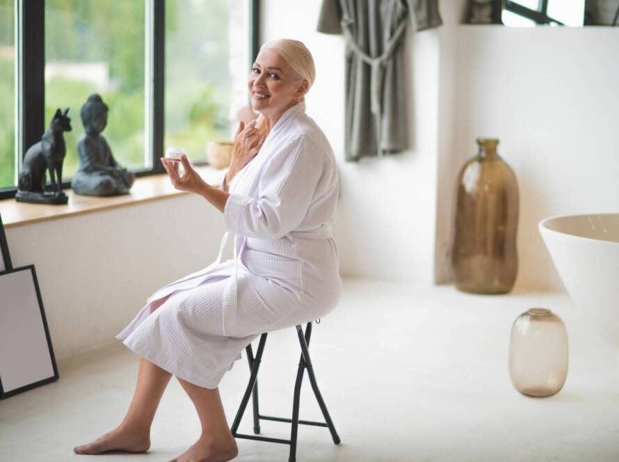An older woman in a dressing gown sits on a stool by a window, applying cream to her face in a modern, minimal bathroom.