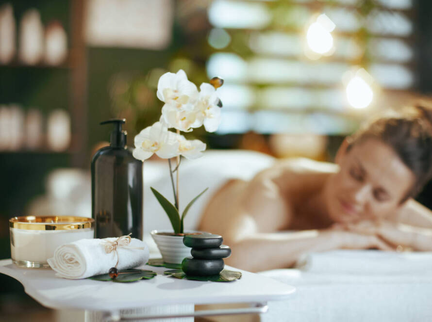 Spa setting with massage stones, a rolled towel, a candle, and a lotion bottle in the foreground; a person is lying on a massage table in the background.