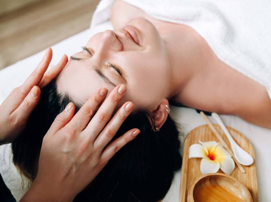 Person lying on a massage table receiving a head massage, with a wooden tray holding a skincare bowl, spoons, and a frangipani flower nearby.