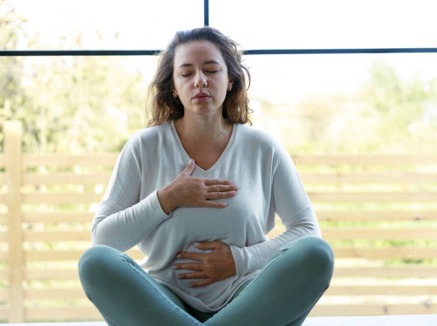 Woman sitting cross-legged indoors with eyes closed, one hand on her chest and one on her abdomen, practising deep breathing or meditation.
