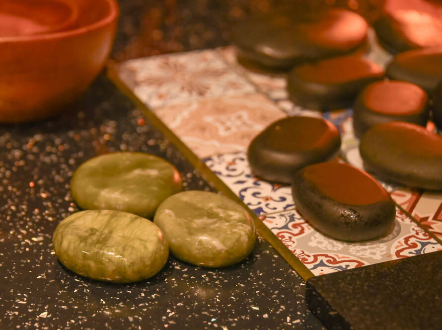 Two green massage stones rest on a black worktop beside several black stones arranged on patterned tiles, with a wooden bowl nearby.