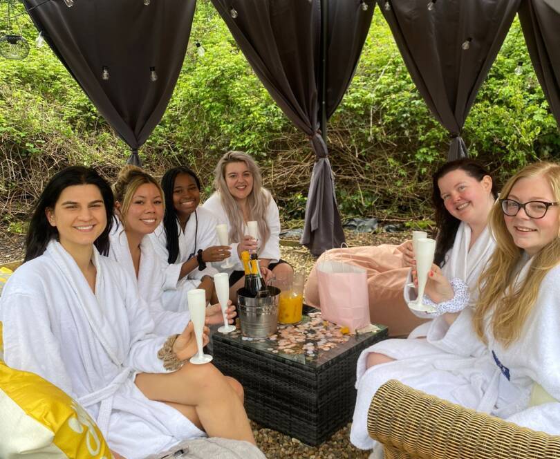 Day spa Surrey UK Six women in white robes sit together under a canopy, holding drinks and smiling, with refreshments and gift bags on a small table in front of them.