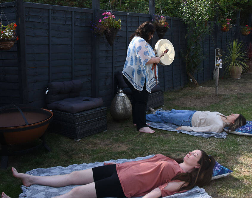 Two women lie on blankets in a garden whilst a third woman stands nearby, holding and playing a drum.