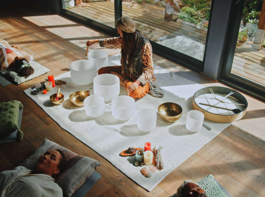 A person sits on a mat playing crystal singing bowls, surrounded by candles and objects, while four others lie down nearby in a bright, indoor space.