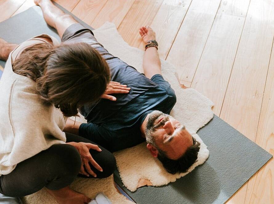 A person lying on a yoga mat with a towel under their head receives assistance with a stretching or relaxation exercise from another individual.