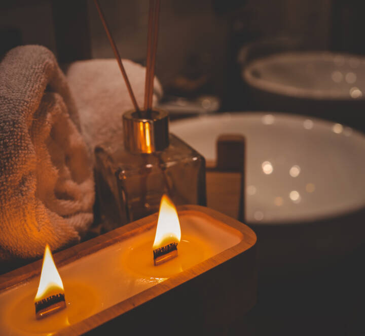 A rectangular candle with two flames sits beside a rolled towel, a glass reed diffuser, and a white basin in a dimly lit bathroom.