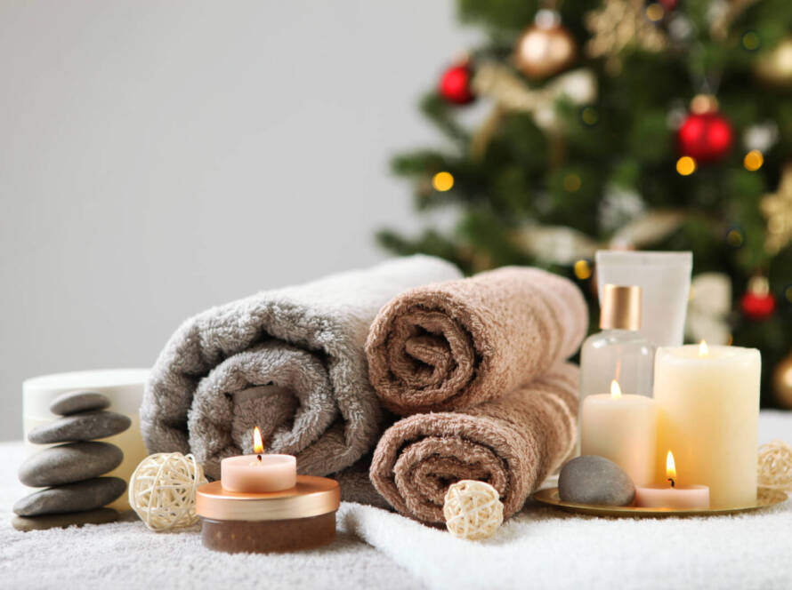 Rolled towels, lit candles, stones, and spa products are arranged on a table with a decorated Christmas tree in the background.