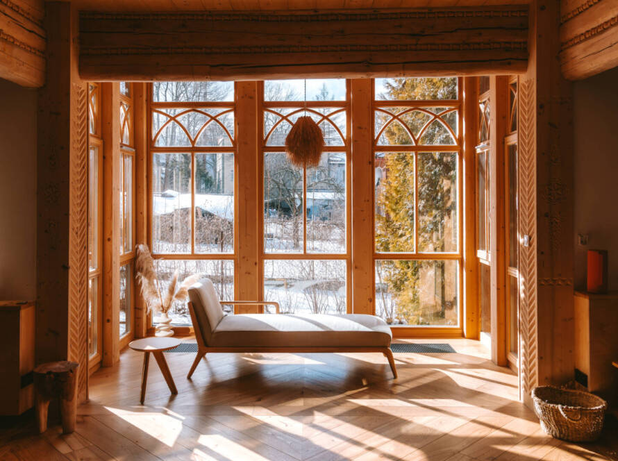 A wooden conservatory with large windows features a chaise longue, small side table, and basket, with sunlight streaming in and a snowy landscape visible outside.