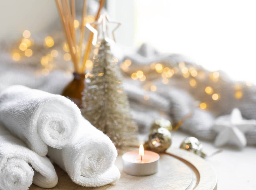 Christmas and New Year Wellness and Rolled white towels, a lit tealight candle, a small decorative tree, and festive baubles are arranged on a tray with warm fairy lights in the background.