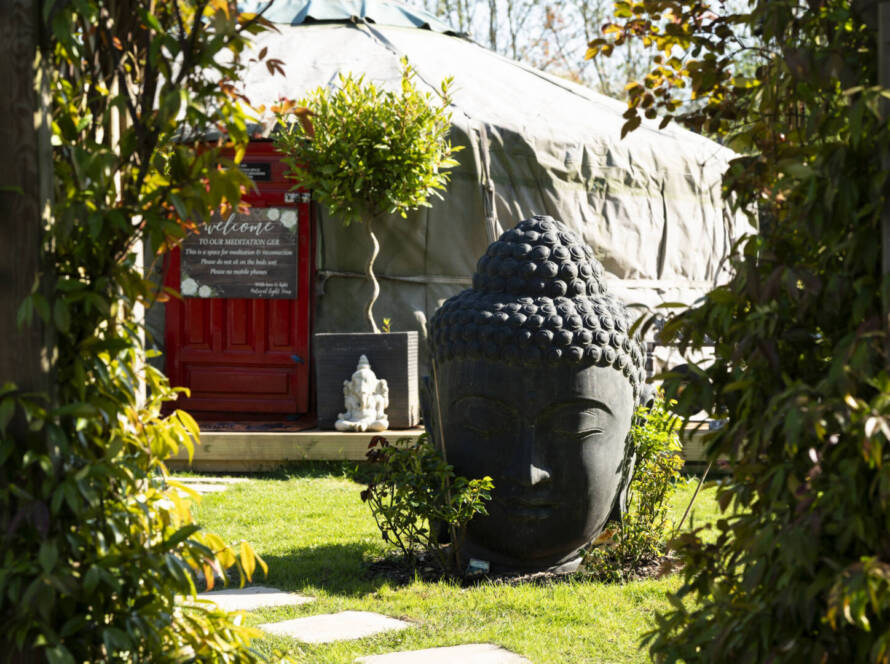 A large stone Buddha head statue is placed on grass in a garden, framed by greenery, near a red door and a round tent structure in the background.