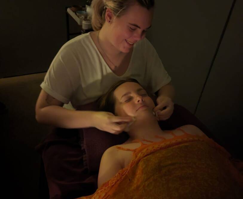 A person is giving a facial treatment to a client lying on a massage table covered with an orange patterned blanket in a dimly lit room.