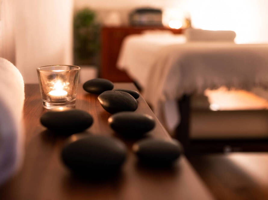 A lit candle and black massage stones on a wooden table with a massage bed and towels in a softly lit spa room.