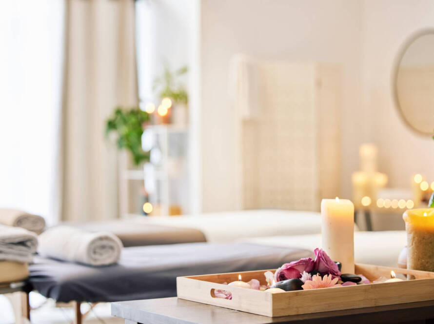 A tray with lit candles, flowers, and stones sits on a table in a softly lit spa room with massage tables and towels in the background.