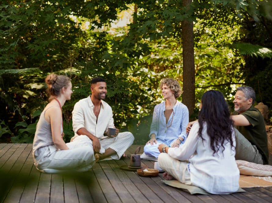 Five people sit on a wooden terrace outdoors, talking and drinking drinks, surrounded by green trees and plants.