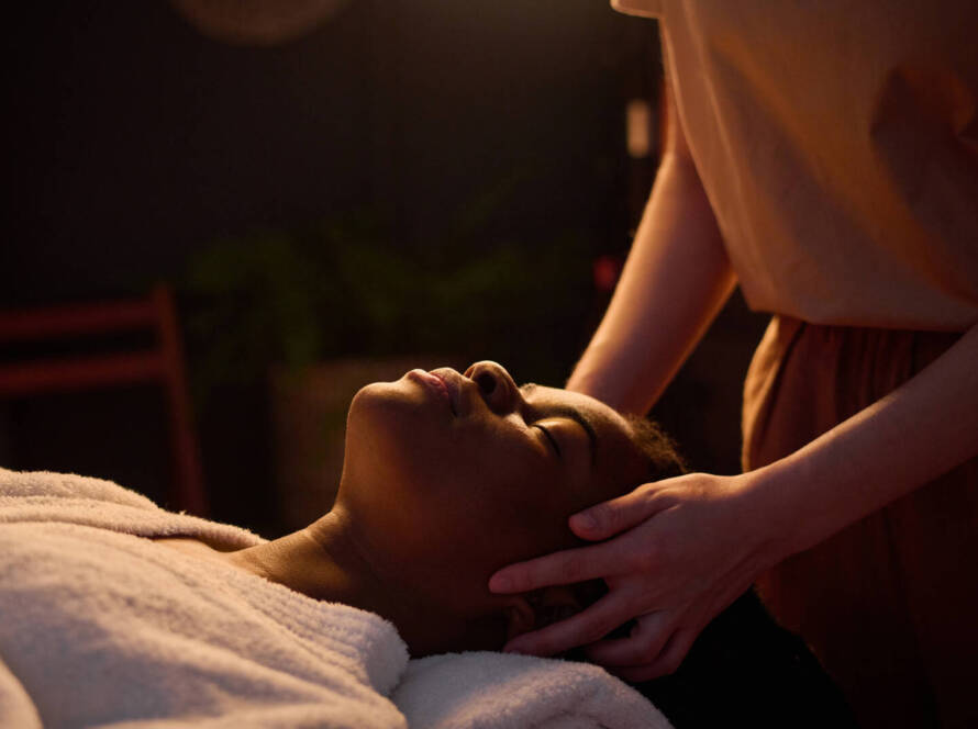 Person lying down with eyes closed, covered by a towel, receiving a head massage from a person standing beside them in a dimly lit room.