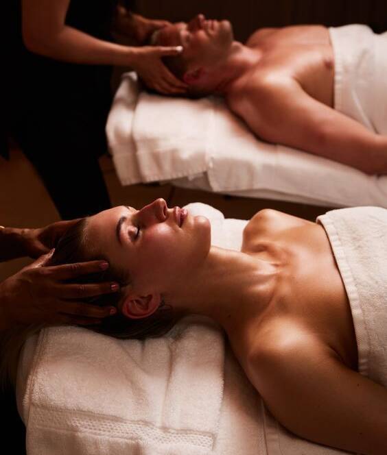 A man and a woman lie on separate spa beds, covered with towels, receiving head massages from two therapists in a dimly lit room.