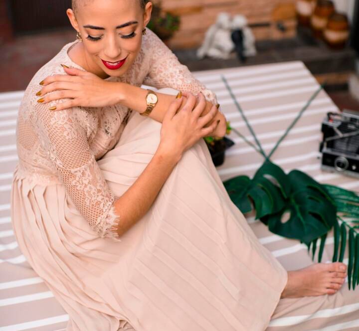 A woman with a shaved head wearing a long, light-coloured dress sits barefoot on a striped surface, smiling and looking down, with plants and a camera nearby.