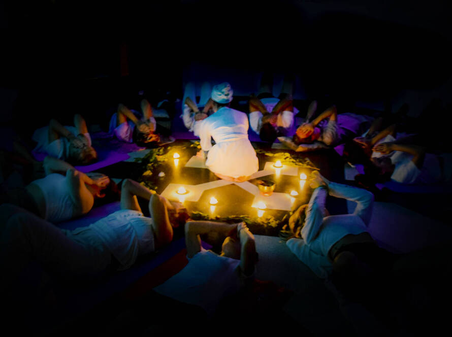 A group of people in white clothing lie in a circle around candles arranged in the centre, with one person kneeling in the middle, in a dimly lit room.