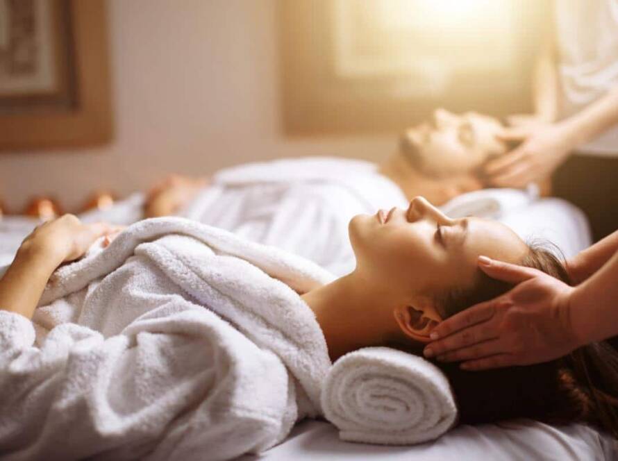Two people lying on massage tables in a spa, wearing white robes, receive head and neck massages from therapists in a softly lit room.