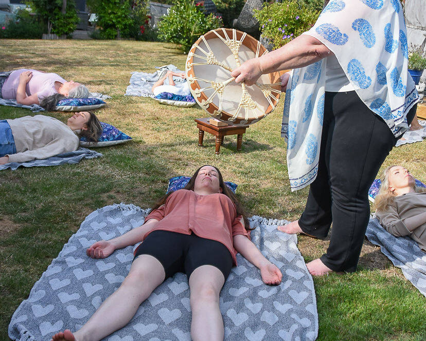 A group of people lie on blankets outdoors whilst a woman stands nearby, holding and playing a large drum.
