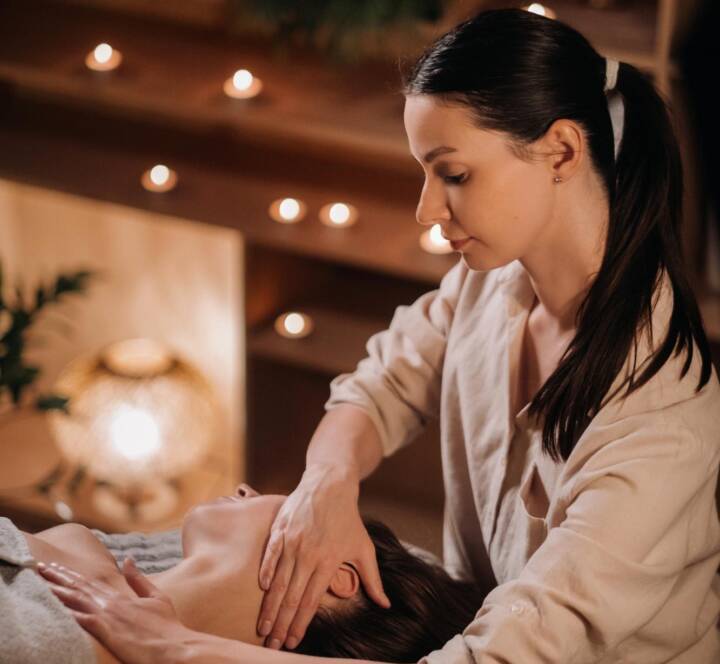 A woman is giving a neck and shoulder massage to a person lying down in a dimly lit room with candles.