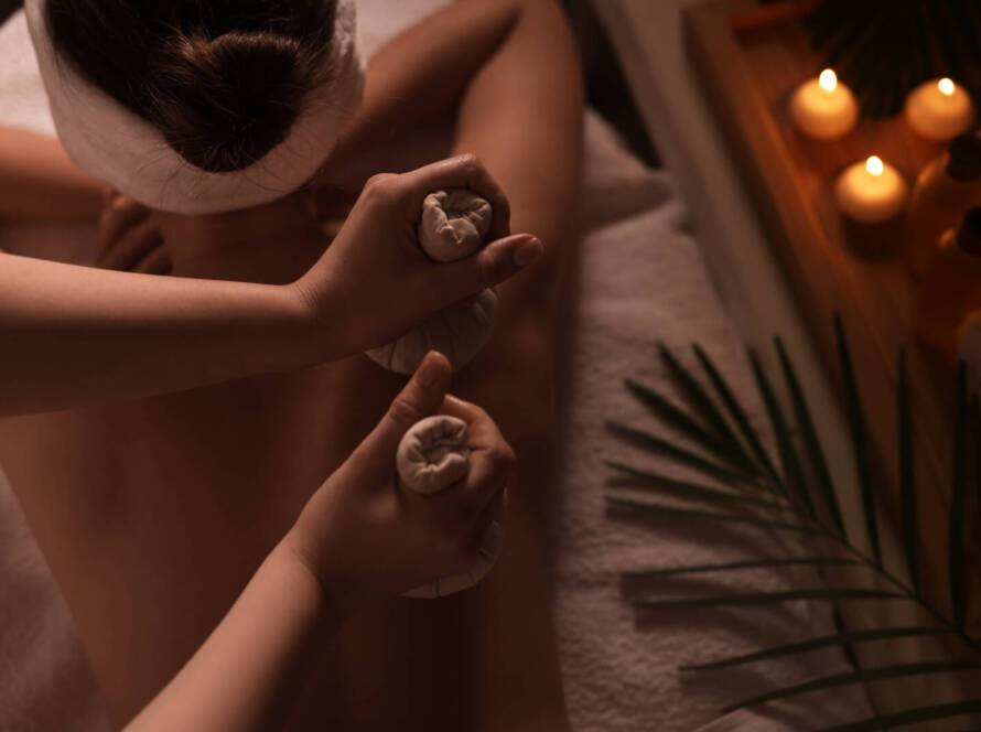 Person receiving a herbal compress massage in a spa setting, with candles, massage oil, and a palm leaf on a tray nearby.