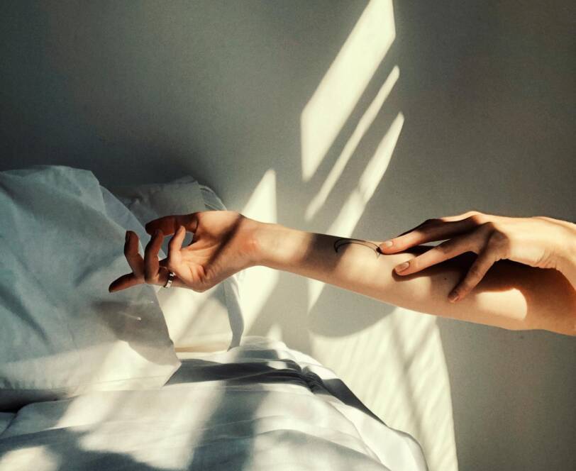 A person’s arm with a small tattoo is outstretched above a white bed, with sunlight casting shadows on the wall and bedclothes.
