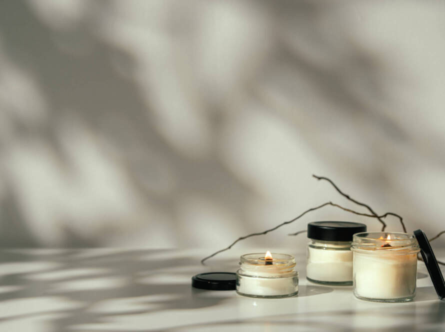 Three small glass jar candles, two lit and one unlit, are placed on a white surface with their lids nearby. A bare twig lies behind them and there are soft shadows on the background wall.