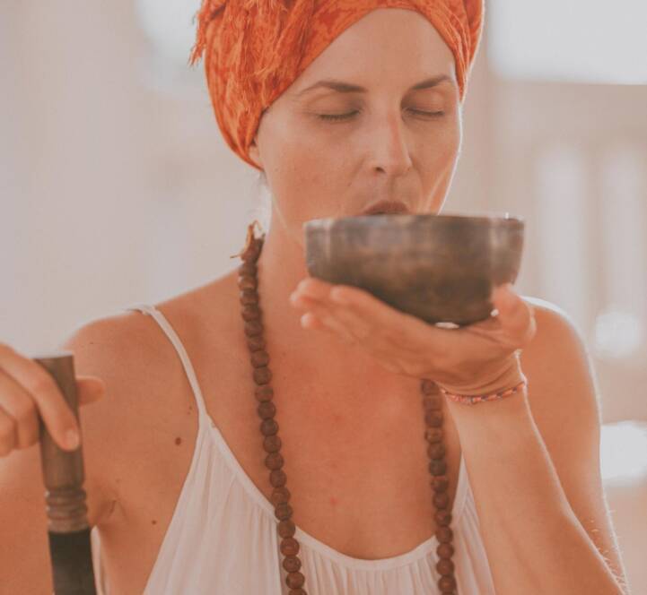 A woman wearing an orange headscarf and beaded necklace holds a singing bowl and mallet, with eyes closed, appearing to meditate or focus on the bowl.