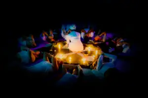 A group of people in white clothing lie in a circle around candles, with one person kneeling in the centre, practising corporate rewilding in wellbeing programmes in a dimly lit setting.