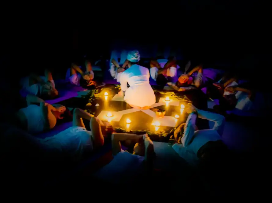 A group of people in white clothing lie in a circle around candles, with one person kneeling in the centre, practising corporate rewilding in wellbeing programmes in a dimly lit setting.