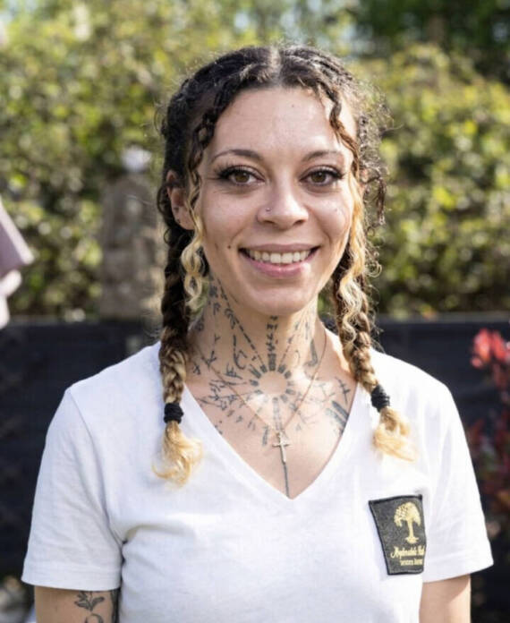 A smiling woman with plaited hair, neck and chest tattoos, a cross necklace, and a white branded T-shirt, photographed outdoors.