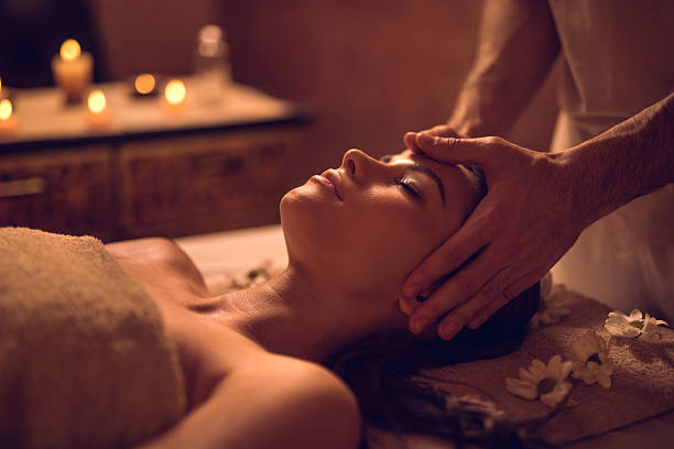 A person lying on a massage table receives a head massage in a dimly lit room with candles and decorative flowers nearby.