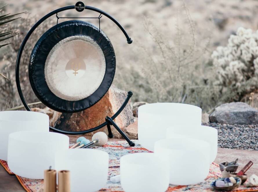 A set of white crystal singing bowls, mallets, a large gong on a stand, candles, and crystals are arranged on a patterned rug outdoors in a desert setting.