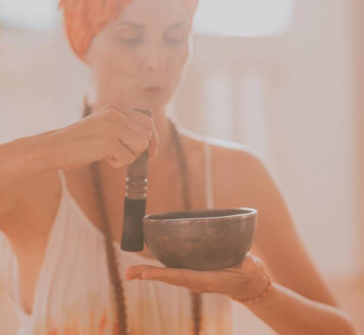 A person wearing an orange headscarf and beaded necklace holds a singing bowl in one hand and strikes it with a beater.
