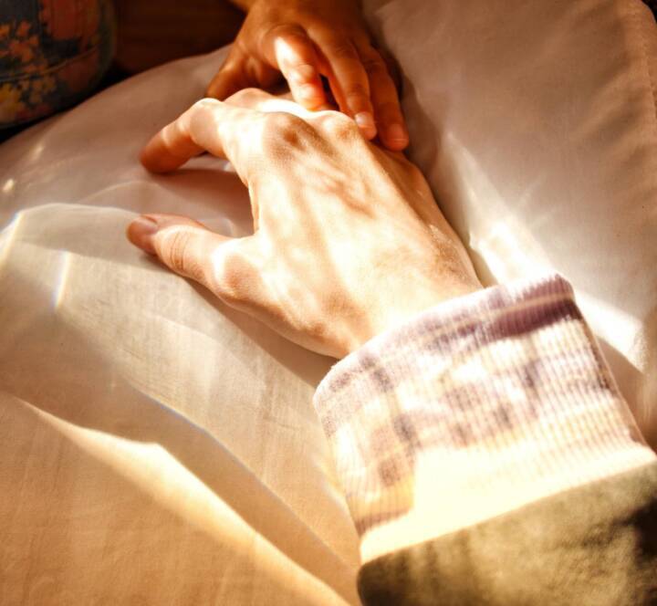 A child’s hand gently resting on top of an adult’s hand on a pillow, with soft sunlight streaming over them.