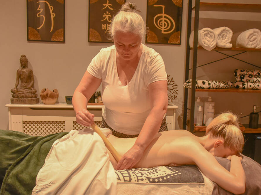 A massage therapist gives a back massage to a client lying face down on a table in a spa room decorated with towels and artwork.