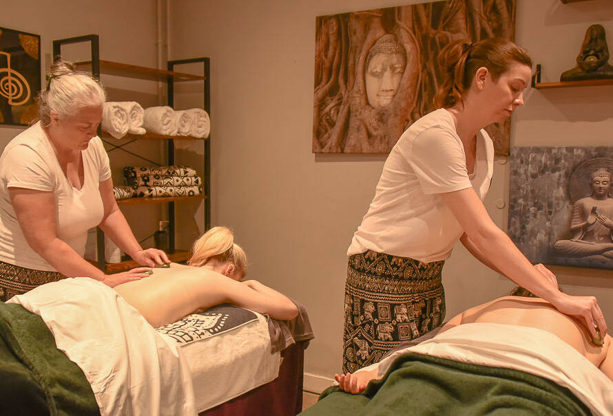 Two massage therapists give massages to clients lying face down on tables in a spa room decorated with Buddha artwork and rolled towels.